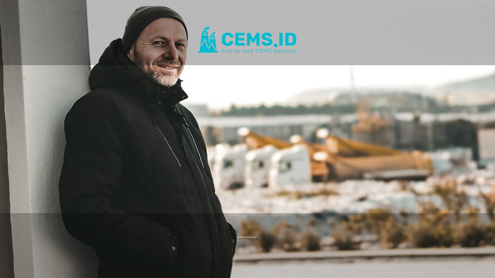 Smiling man in winter attire outdoors leaning against a wall with snowy background.