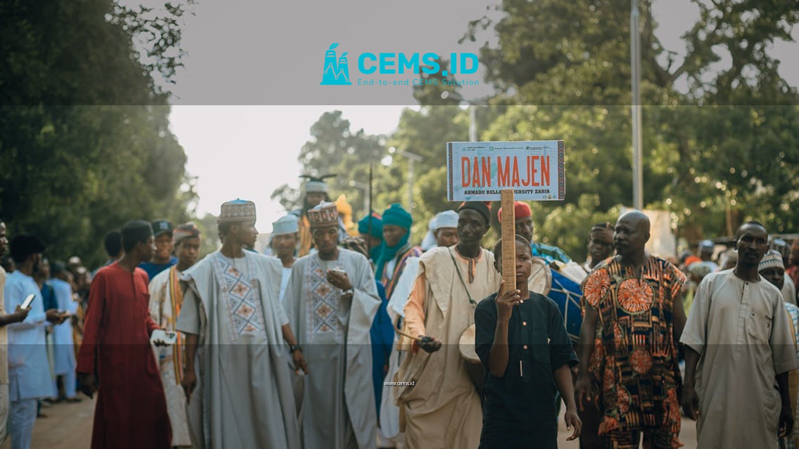 A vibrant cultural procession with a diverse group holding a 'Dan Majen' sign outdoors.