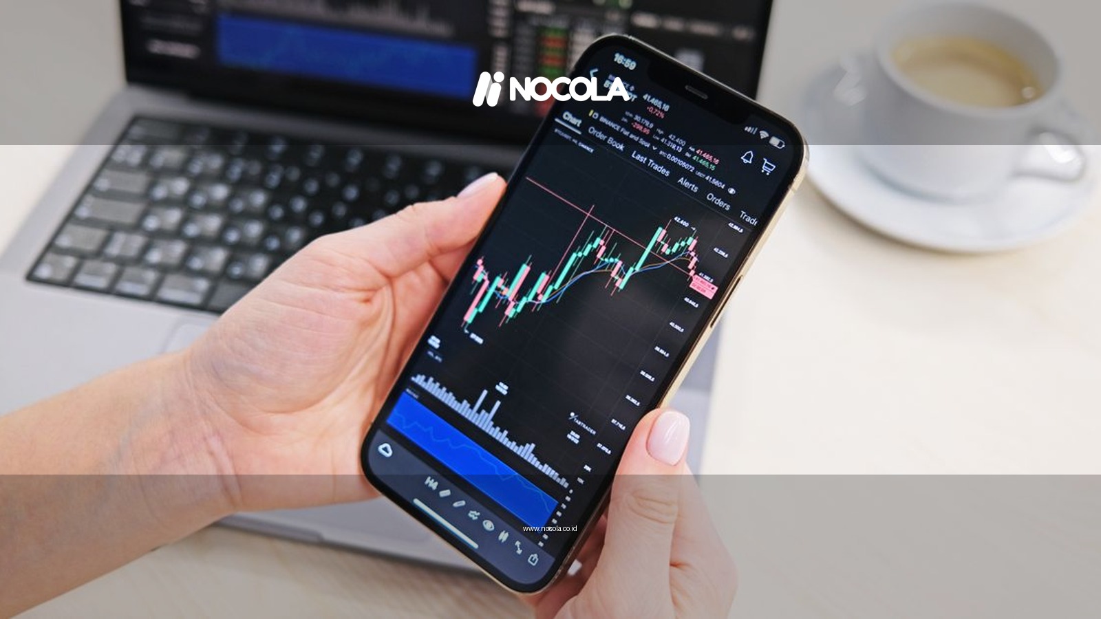 Close-up of hands holding smartphone displaying stock charts with laptop in the background on a desk.