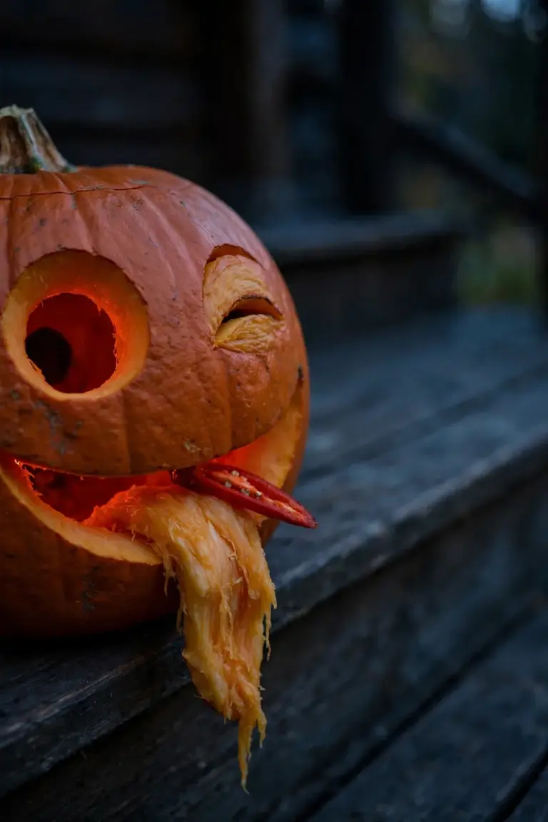 Winking carved pumpkin with one wide round eye, a lopsided grin and a red tongue hanging from its mouth