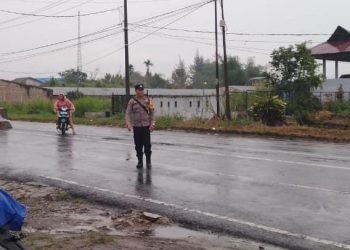 Bhabinkamtibmas Polsek Raya, Aipda Joni Susanto Purba, tetap menjalankan tugasnya mengatur lalu lintas, di Simpang Sekolah SD Bintang Timur, pada Jumat (14/03/2025) pagi.